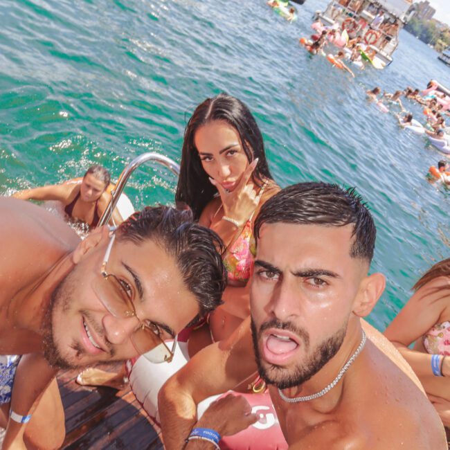 Three young adults in swimsuits pose on a boat’s deck, smiling and making playful faces. Other people are swimming and relaxing on boats in the blue water under a sunny sky. "Sail Gustavo" logo is in the corner.
