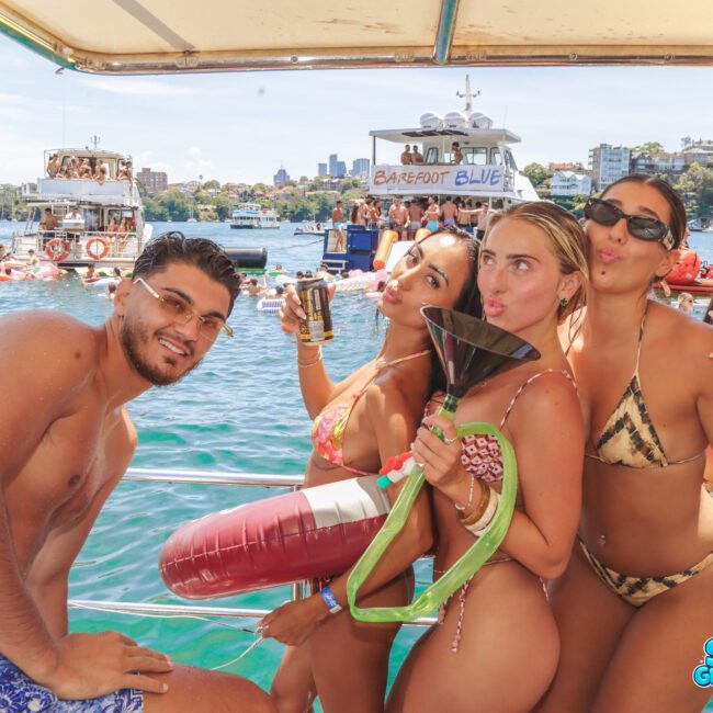 A group of four young adults in swimwear pose and smile on a boat, holding drinks and a beverage bong, with water, inflatables, and other boats in the background. The atmosphere is lively and festive.