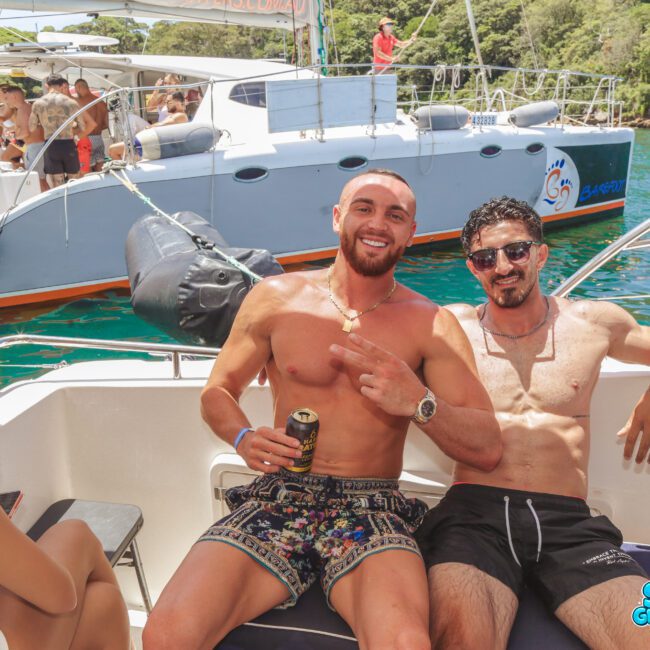 Two smiling men in swimwear sit on a boat, enjoying drinks and posing for the camera. A group of people and another boat are visible in the background, with trees and water surrounding the scene.