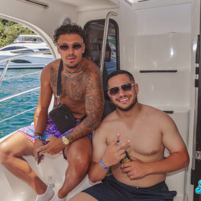 Two smiling men sit shirtless on a boat, one with tattoos and sunglasses, the other making a peace sign. The background shows water, a yacht, and green trees. Blue "Sail Gustavo" logo is in the corner.