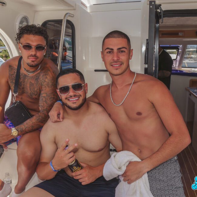 Three young men in swimwear pose and smile on a boat. Two sit while one stands behind them with his arm around a friend. The scene is bright and sunny, with water and other boats in the background.