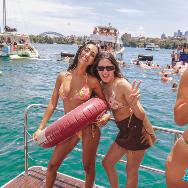 Two women in swimsuits smile and pose on a boat, with one holding a maroon float. Other people and boats are visible in the background on the water, with city buildings and the Sydney Harbour Bridge in the distance.