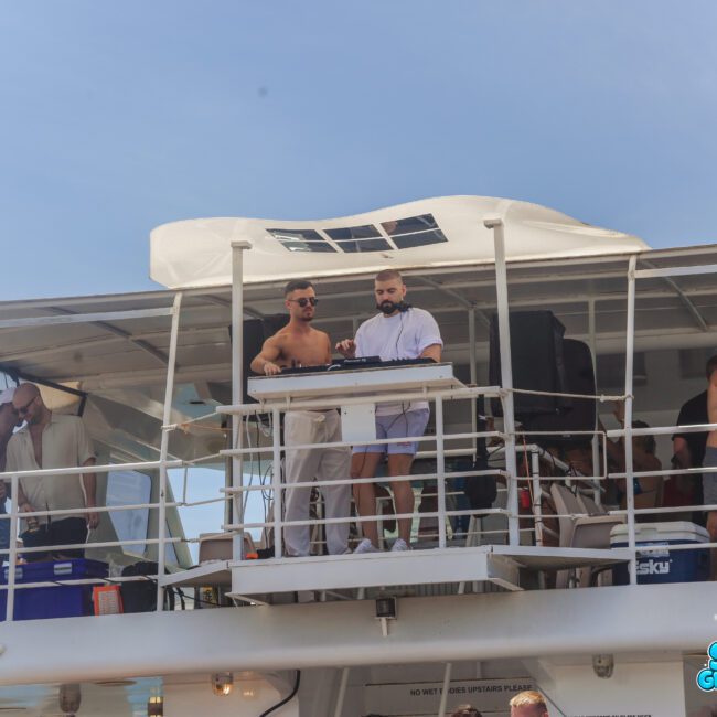 A group of people enjoy a party on a boat. Two men stand at a DJ booth on the upper deck, one wearing white and the other holding headphones. Others lounge and socialize around them under the sunny sky.