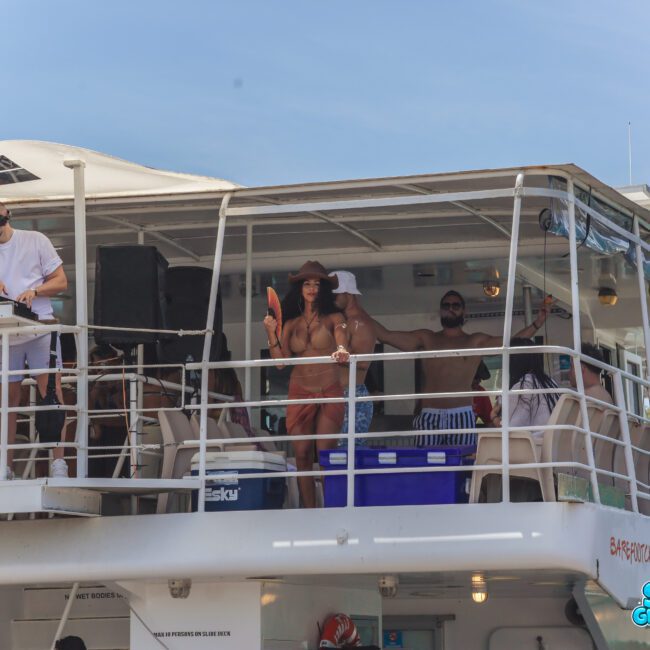 A group of people enjoy music and dancing on the upper deck of a white boat under a clear blue sky. Some are wearing swimsuits, sunglasses, and hats. A DJ is performing on the left side of the deck.