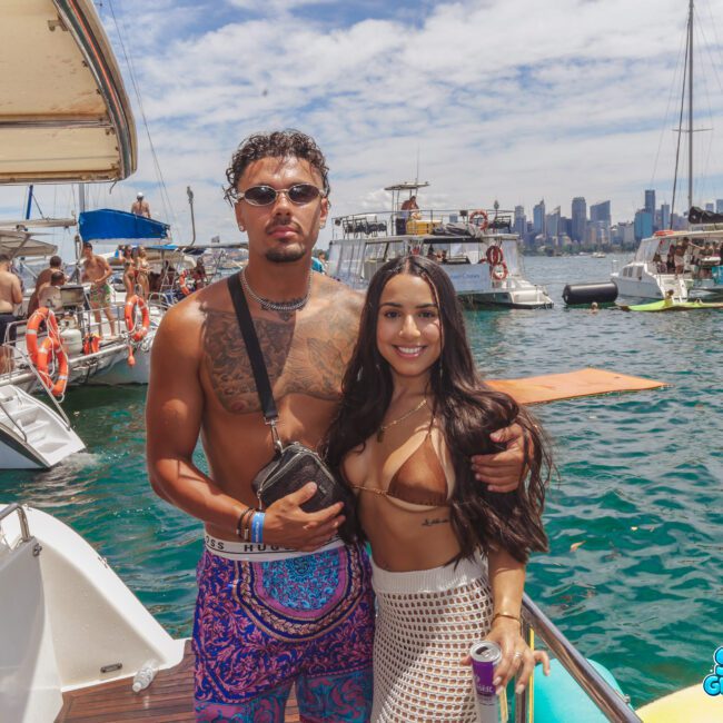 A man and woman pose together on a boat with several other boats and people in the background. The man is shirtless and wears sunglasses, while the woman wears a bikini top and crochet pants. The city skyline is visible in the distance.
