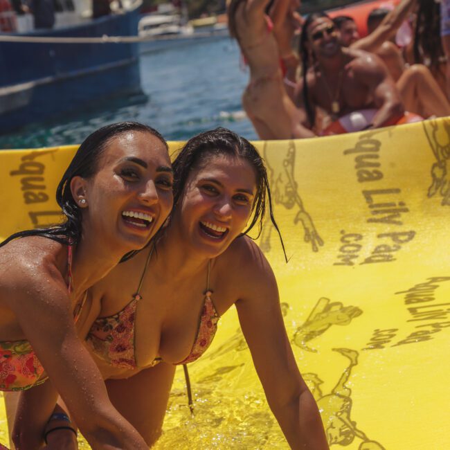 Two smiling women in swimsuits sliding on a yellow inflatable water slide, with others in swimwear cheering in the background near a boat and water under sunny skies.