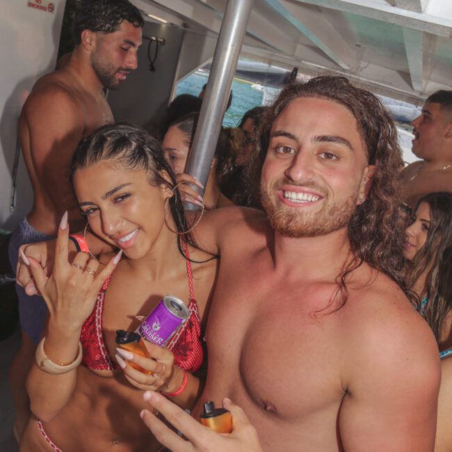 A group of young adults in swimsuits smiling and posing for the camera on a boat. One woman in a red bikini holds a drink can and makes a hand gesture, while a man next to her smiles and holds a small bottle.