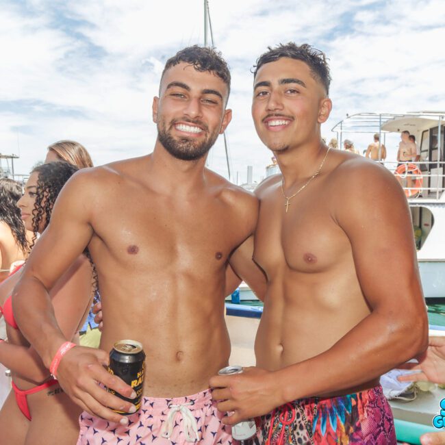 Two young men in swim trunks smile and pose together on a boat, holding drinks. There are other people in swimwear and boats in the background, suggesting a lively beach or party atmosphere.