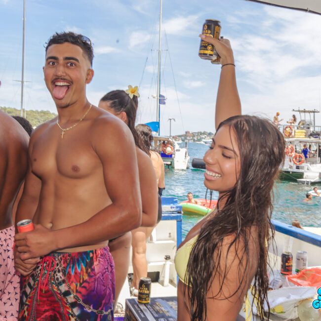 A group of young people on a boat enjoy a sunny day; a shirtless man makes a funny face while a smiling woman with wet hair raises a can. Other boats and people are visible on the water in the background.