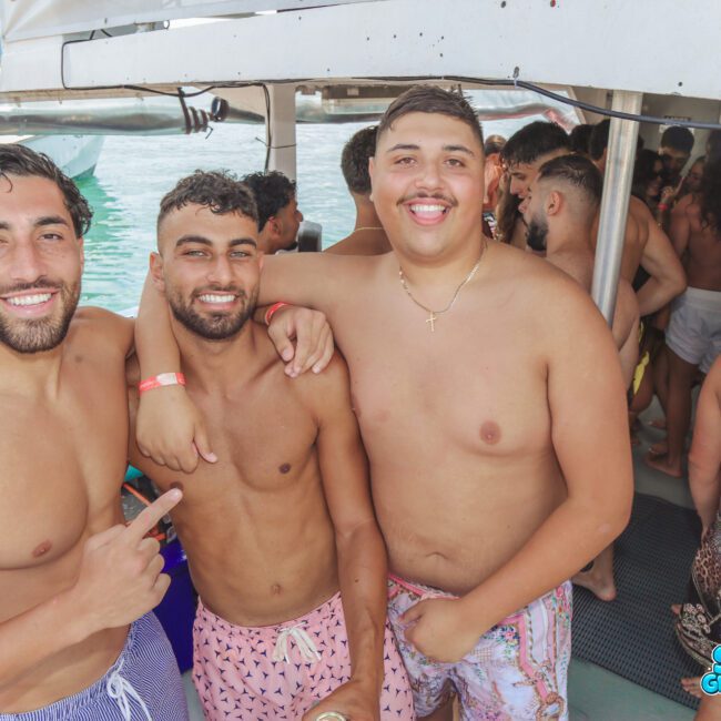 Three smiling men in swim trunks pose together on a boat during a lively party, with other people in the background. The boat is docked near water, and the atmosphere is relaxed and cheerful.