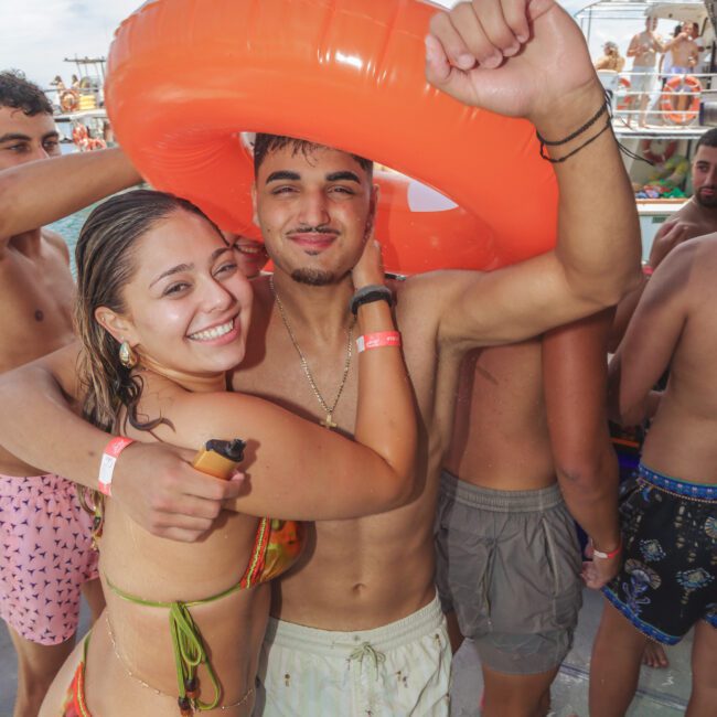 A smiling woman in a bikini hugs a man while they both pose under an orange pool float at a lively pool party on a boat. Other people in swimwear are mingling in the background under a partly cloudy sky.