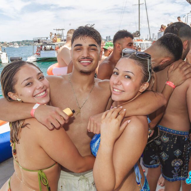A group of smiling young adults in swimsuits enjoy a lively boat party, hugging and posing for a photo with the ocean and other boats in the background.