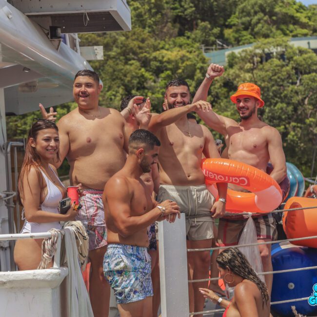 A group of young adults in swimsuits stand and smile on a boat, some holding drinks and inflatables, with trees and water in the background. They appear to be enjoying a sunny day.