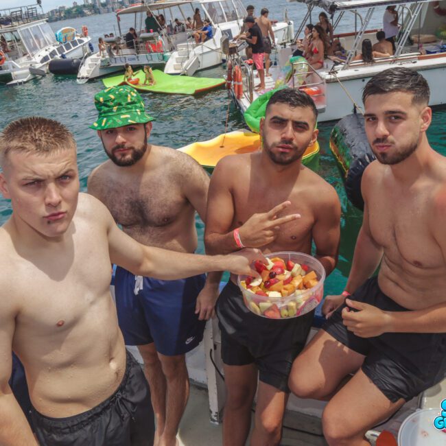 Four young men in swim trunks stand on a boat, holding a bowl of fruit. Behind them are other boats and people enjoying a sunny day on the water. The scene is lively and relaxed.