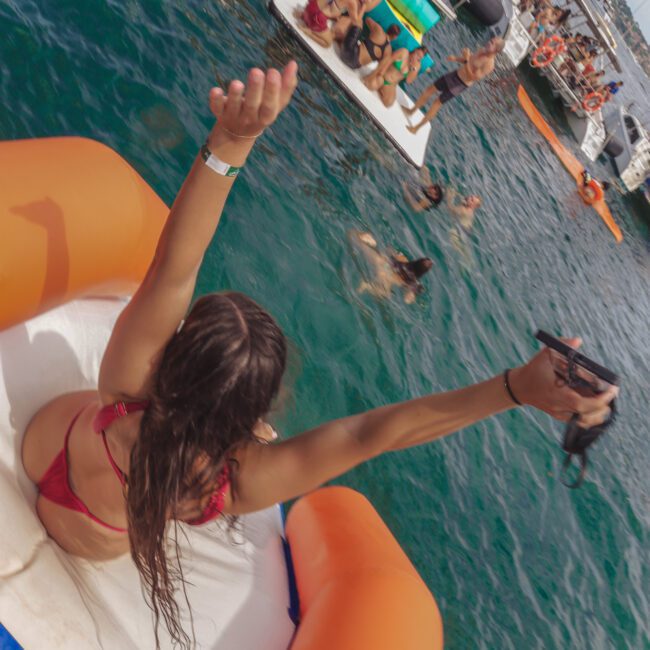 A woman in a red bikini with arms raised slides down an inflatable into the sea, surrounded by people swimming and relaxing on boats during a lively water party.