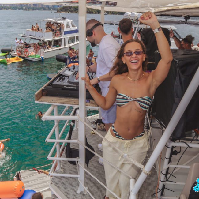 A woman in sunglasses and a striped bikini top dances and smiles on a boat with a DJ setup. Other people are swimming and relaxing on nearby boats in bright blue water. The scene is lively and festive.
