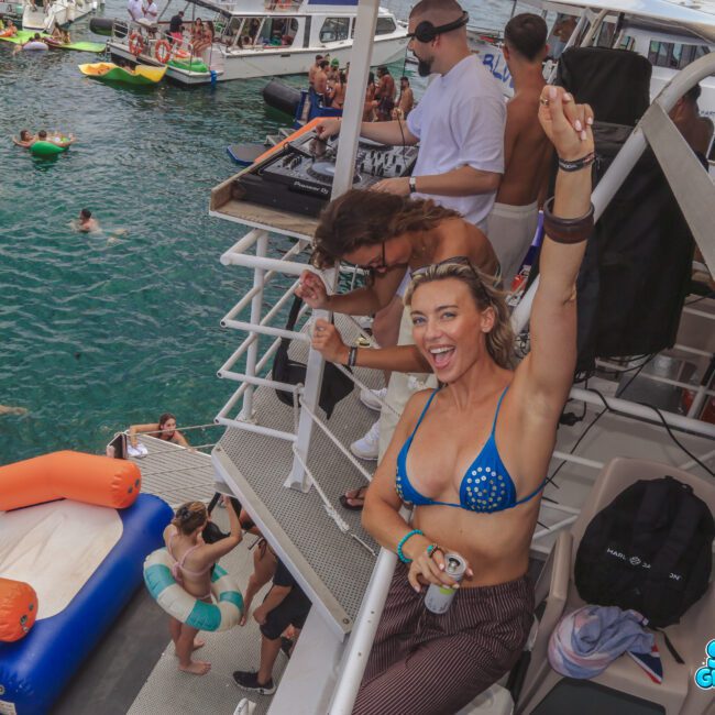 A woman in a blue bikini top smiles and raises her arm in celebration on a crowded boat, surrounded by people enjoying a pool party with boats and inflatable floats in the water. "Sail Gustavo" logo appears in the corner.