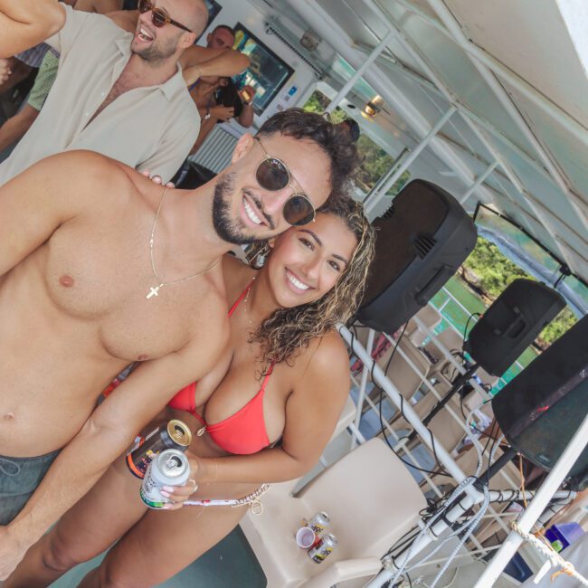 A smiling man and woman in swimwear pose together on a boat party, holding drinks. Other people are mingling in the background. The atmosphere is lively and summery, with music equipment visible behind them.