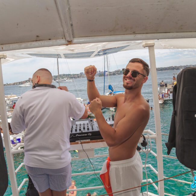 Two men stand on a boat; one is DJing, and the other is shirtless, wearing sunglasses, smiling, and flexing his arms. Boats and people are visible on the blue water in the background.
