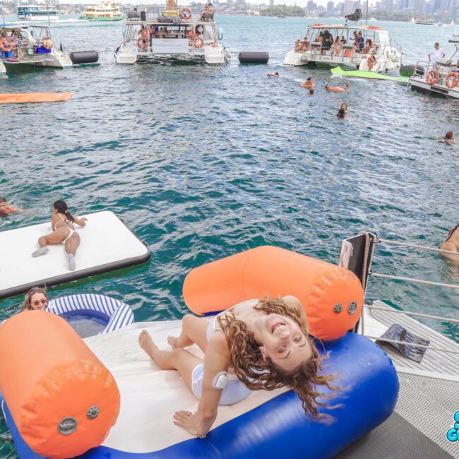 People enjoy a boat party on the water with inflatable floats and rafts. A smiling woman lounges on a water float in the foreground, while others swim and relax nearby. Several boats are anchored in the background.