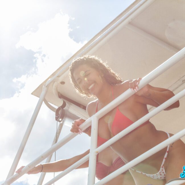 A woman in a bikini smiles and leans over the white railing of a boat under a partly cloudy sky. Sunlight creates a bright, cheerful effect. The "Sail Gustavo" logo appears in the bottom right corner.