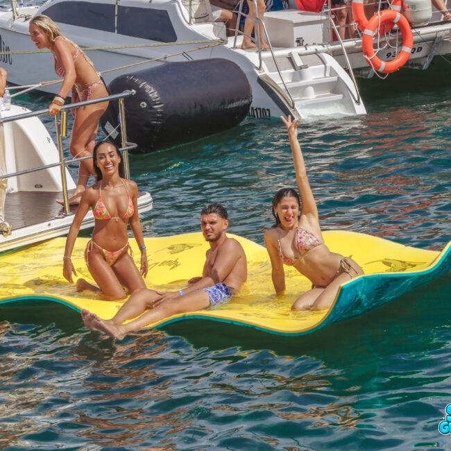 Three young adults in swimsuits relax and smile on a floating yellow mat in the water, next to a boat with other people enjoying the sunny day. The sea is clear and blue.