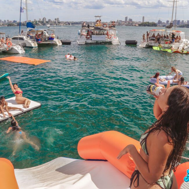 A lively boat party scene with people swimming and relaxing on floats and boats in the water. One woman, holding a drink, stands on an orange and white inflatable. City skyline and blue sky are visible in the background.