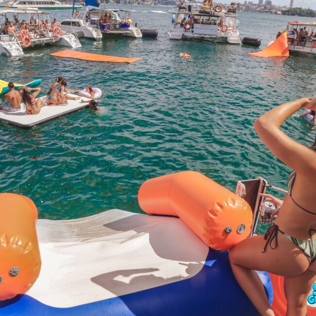 A woman in a bikini sits at the top of an inflatable slide on a boat, looking out at people lounging on floats and swimming in turquoise water near several anchored party boats. The city skyline is visible in the distance.
