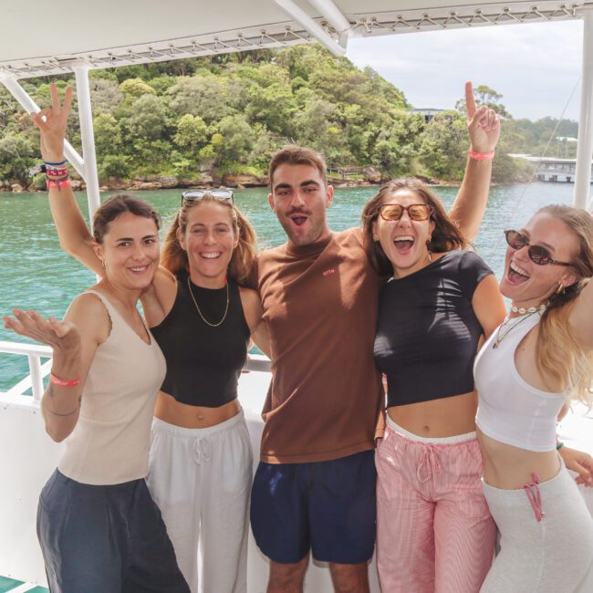 Five friends smile and pose with arms around each other on a boat, with green water and a forested shoreline in the background. It appears to be a sunny day, and other people are visible on the boat behind them.