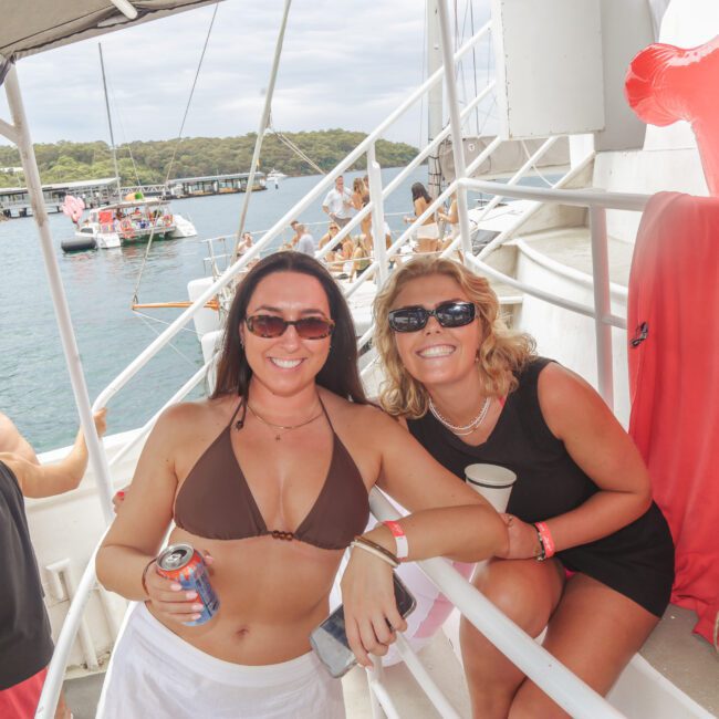 Two women in swimsuits and sunglasses smile at the camera on a boat with drinks in hand. Other people are relaxing nearby. Water, trees, and another boat are visible in the background.