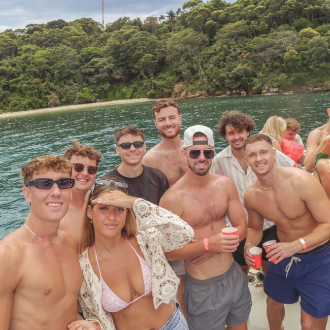 A group of young adults, some shirtless and others in swimwear, smile and pose on a boat with drinks in hand. Behind them is clear blue water and a lush, green forested shoreline.