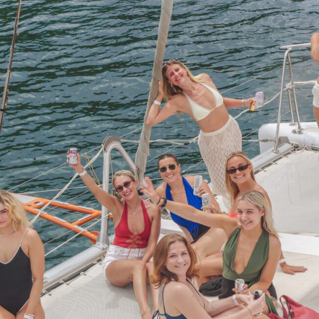 A group of smiling women in swimsuits relax and hold drinks on a sailboat, enjoying a sunny day on the water. Some are sitting, some standing, with the blue water and boat’s deck visible.