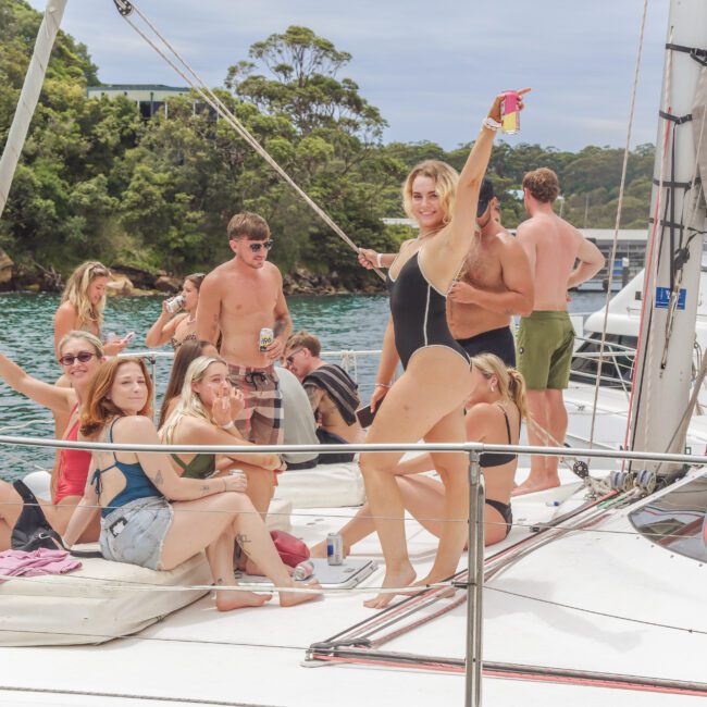 A group of people in swimsuits relax and pose on a sailboat, smiling and enjoying drinks. Trees and water are visible in the background, indicating a fun day out on the water.