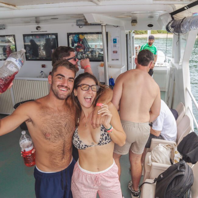 Two smiling people in swimsuits pose arm-in-arm on a boat, with others in the background. The man holds up a large drink bottle. The boat is on water near a wooded shoreline. It's a sunny, lively atmosphere.