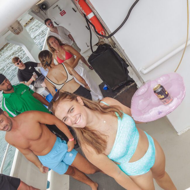 A group of people in swimsuits socializing on a boat. One woman in a blue bikini smiles at the camera while holding a pink inflatable drink holder with a can inside. Other people are chatting and relaxing by the railing.