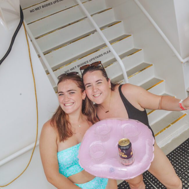 Two women in swimsuits smile at the camera while holding a pink float with drinks. A man in swim trunks holding a can stands nearby on a boat deck. Stairs and safety signs are visible in the background.