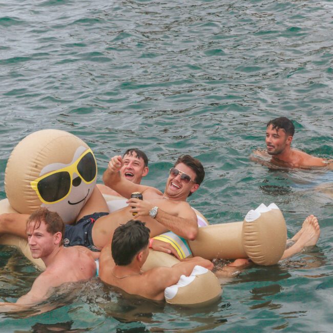 Five young men are enjoying themselves in the water; four are lounging on a large inflatable shaped like a sloth wearing sunglasses, while one person floats nearby. The mood is playful and relaxed.