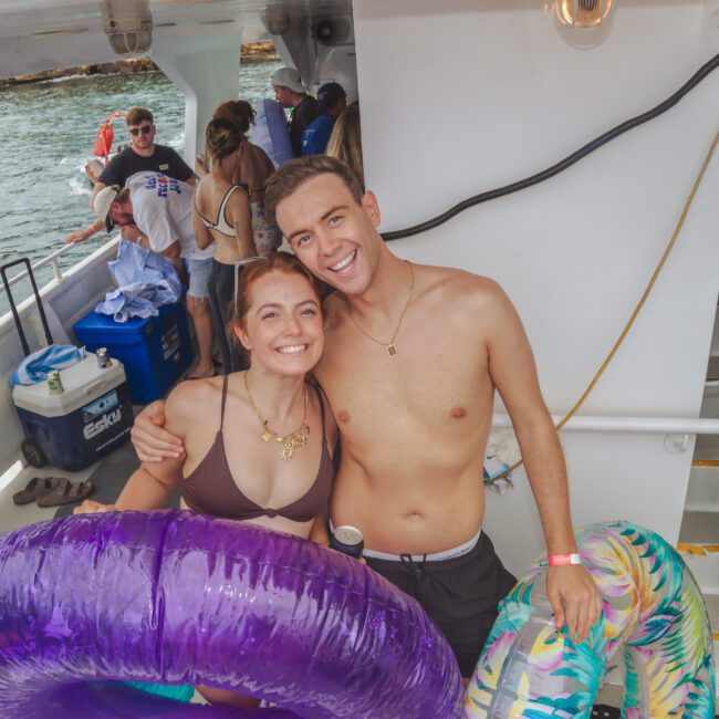 Two people in swimsuits smile and pose together on a boat, each holding inflatable pool floats. Other people are in the background, with some sitting and standing. The scene looks like a fun boat party by the water.