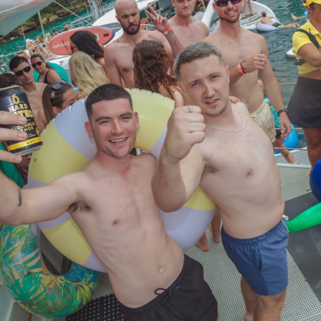 A group of people partying on a boat; two shirtless men pose for a selfie at the front, one with a drink, inflatable tube, and thumbs up. Others mingle in swimwear near clear blue water.