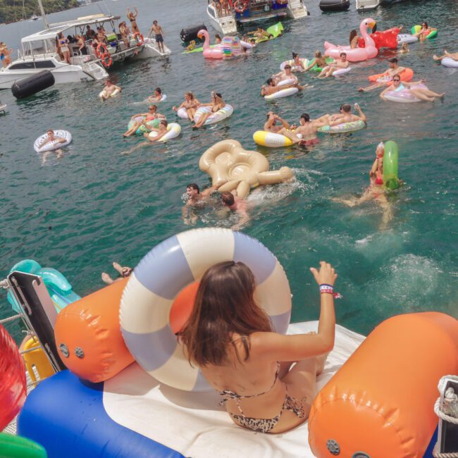 A woman on an inflatable slide descends into a lake crowded with people on colorful pool floats, surrounded by boats on a sunny day. The scene is lively and festive with many enjoying the water.
