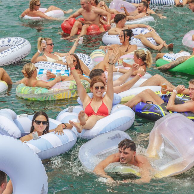 A large group of people relax and have fun on colorful inflatable tubes in a crowded pool or lake, some with arms raised and drinks in hand, enjoying a sunny day.