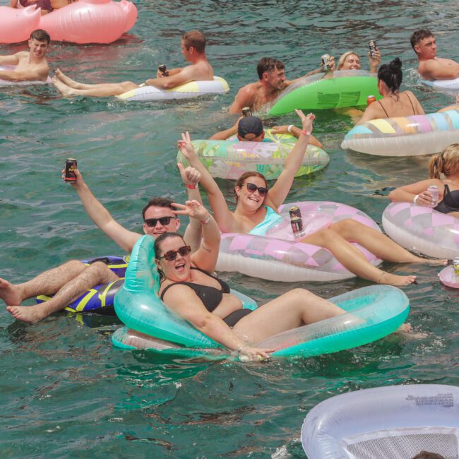 A group of people relaxing on colorful inflatable pool floats in clear blue water, smiling and making peace signs. Some are holding drinks, enjoying a sunny day with friends.