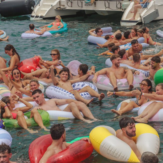 A large group of people relax and have fun on colorful inflatable pool floats in the water near boats, enjoying a lively summer party under sunny weather.