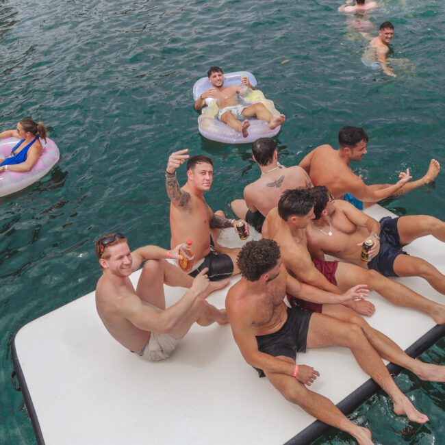 A group of young people relax and socialize on floating inflatables in a pool. Several men sit together on a large white float, smiling and taking selfies, while others float nearby on colorful inner tubes.