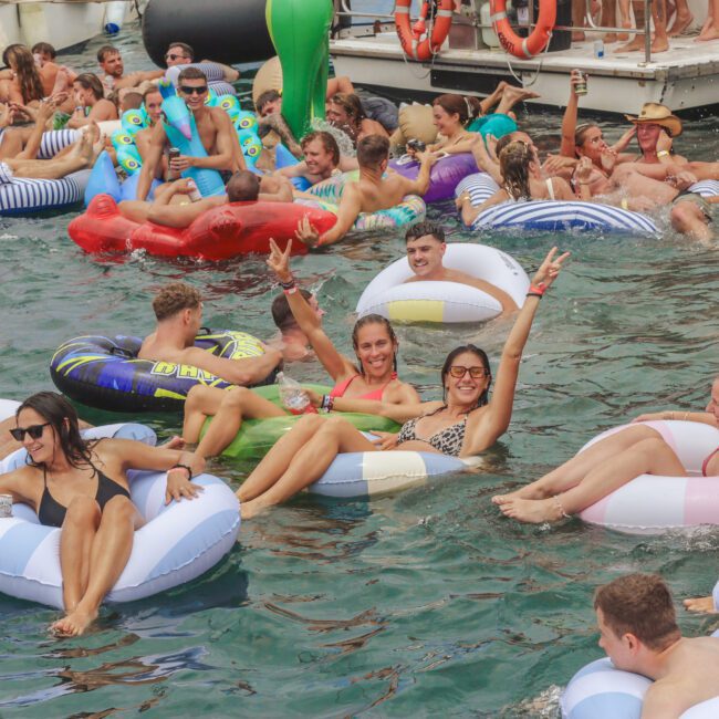 A large group of people in colorful inflatable floats relax and celebrate in the water near several boats, smiling, chatting, and making peace signs on a sunny day.