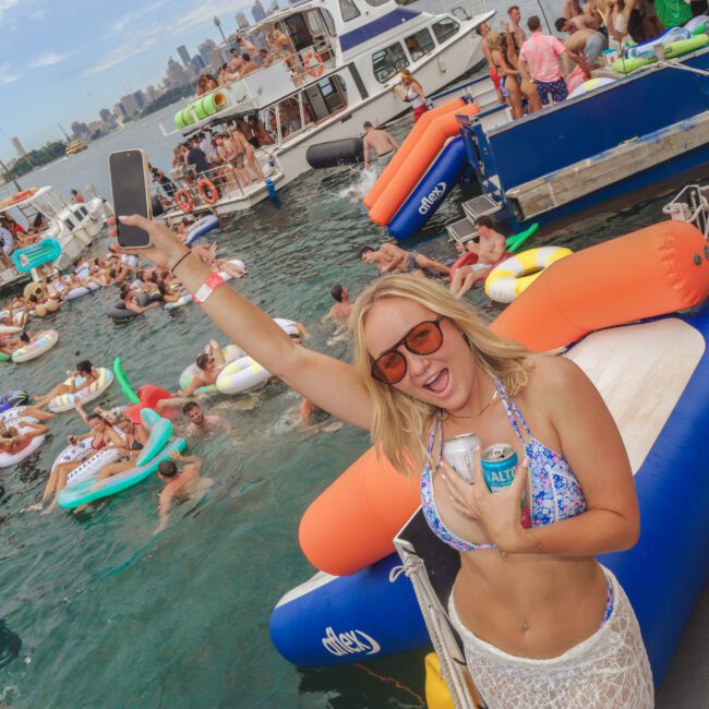 A woman in sunglasses smiles and raises her phone at a lively boat party with people on inflatables and boats in the water, city skyline visible in the background. She holds a drink and wears a swimsuit and crocheted cover-up.