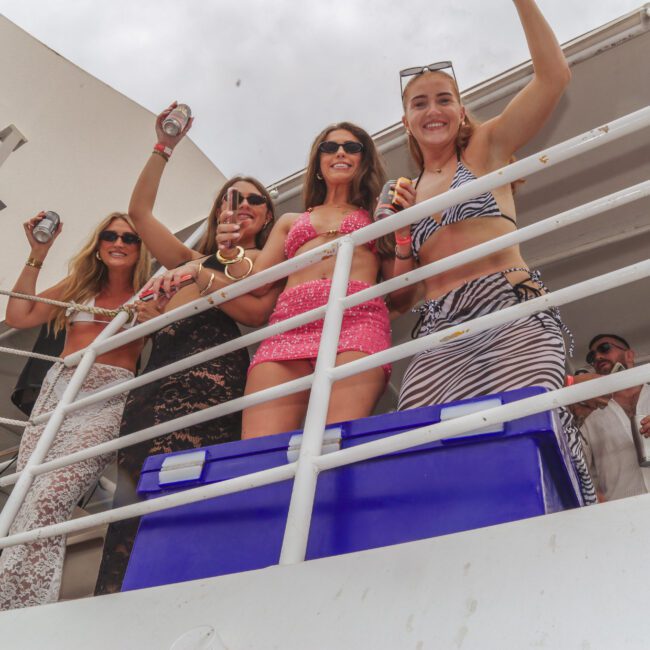 Four women smile and pose on the deck of a boat, holding drinks and wearing sunglasses and summer outfits. The sky is cloudy and a blue cooler is in front of them.