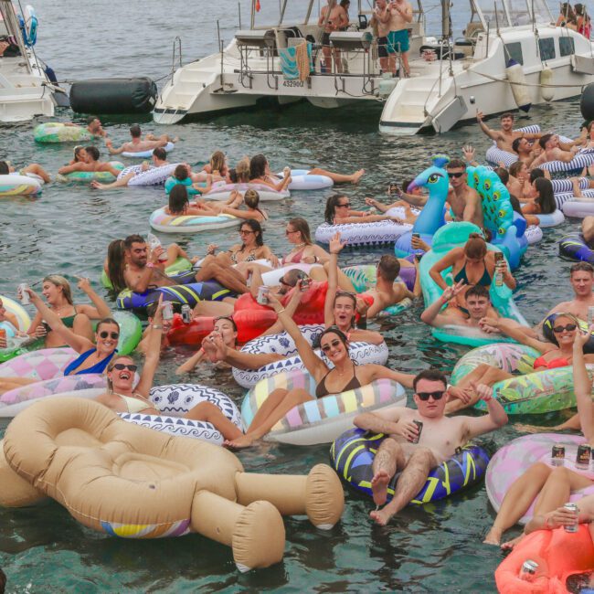 A large group of people float on colorful inflatables in the water near boats, enjoying a lively pool party under clear skies. Everyone appears relaxed and happy, surrounded by a festive summer atmosphere.