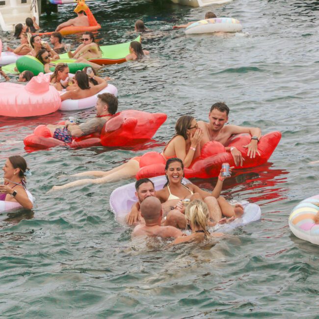 A group of people relax on colorful pool floats in a lake, chatting, laughing, and enjoying drinks. The floats include swans, flamingos, and donut shapes, creating a lively, fun summer atmosphere.