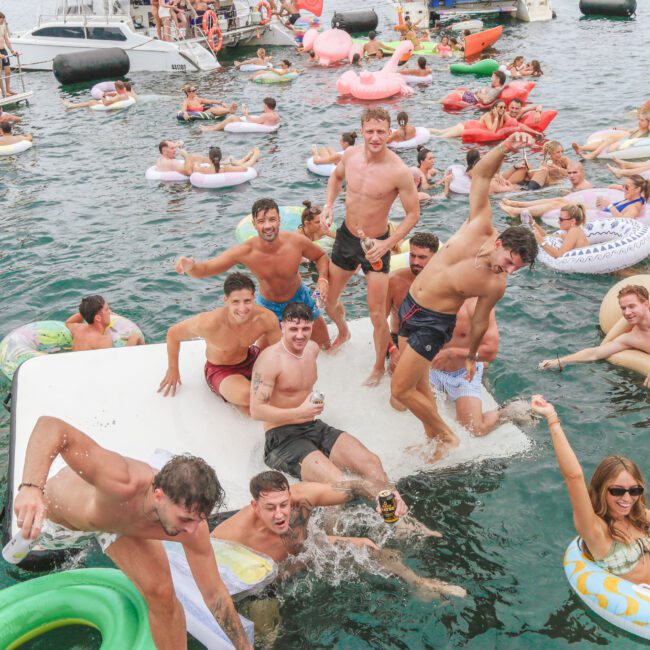 A large group of people in swimsuits enjoy a lively party on the water, lounging on colorful inflatables and dancing on a floating platform, surrounded by boats on a sunny day.